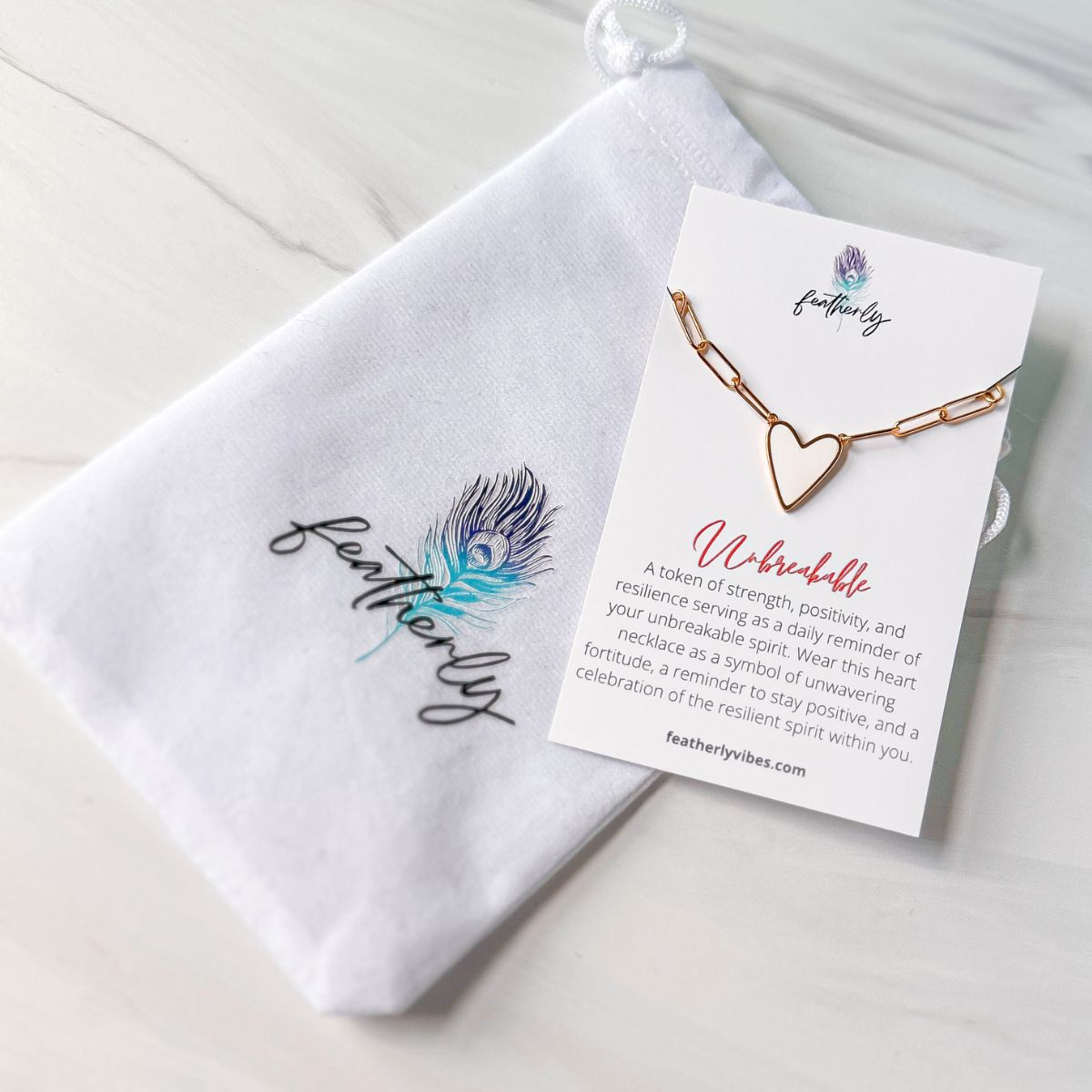 A white enamel heart paperclip chain necklace in gold on the unbreakable meaning card with the Featherly white velvet pouch